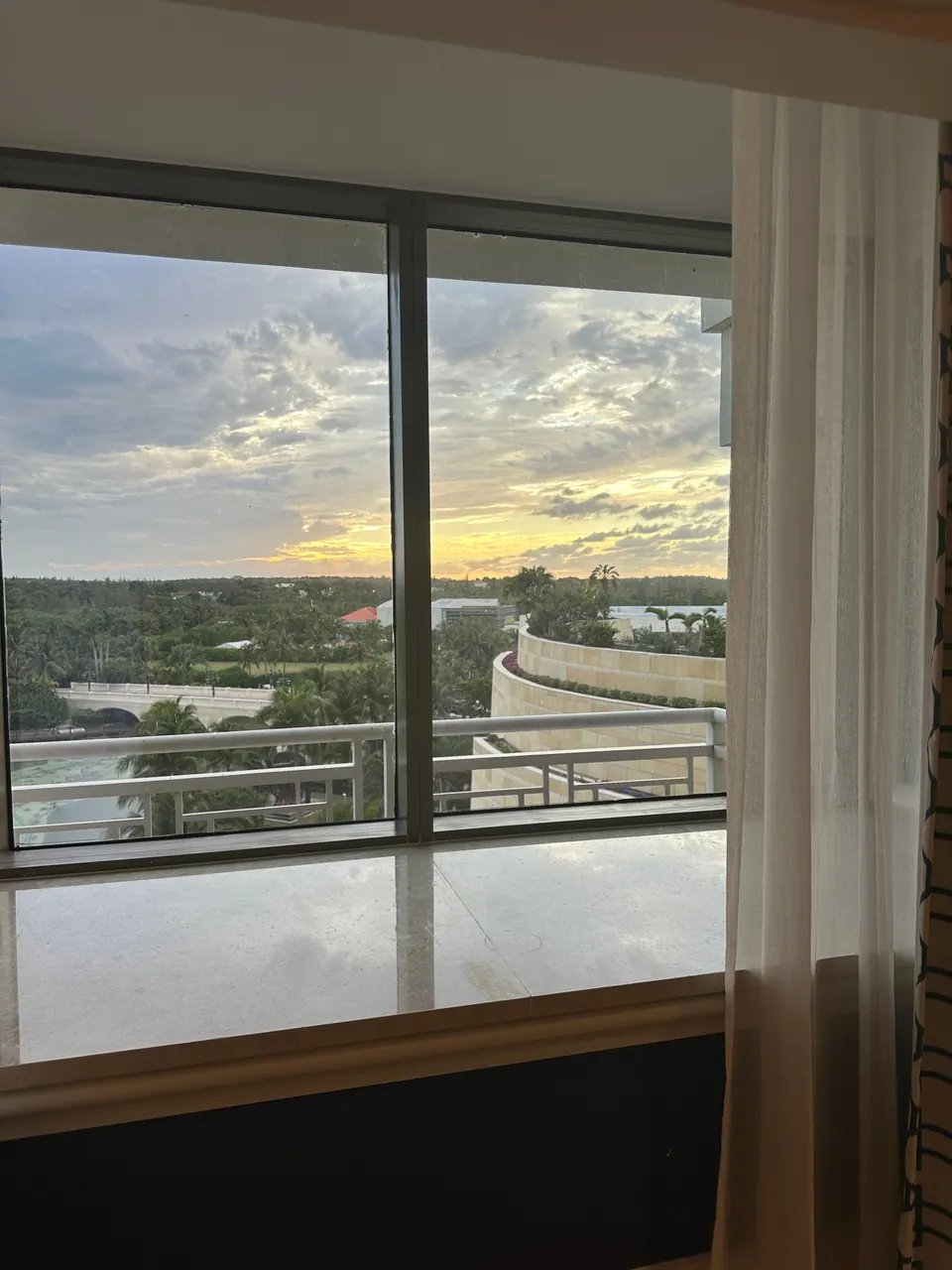 Through a hotel room's floor-to-ceiling window, a cloudy sunset glows over a sea of treetops, a curved balcony and the resort grounds.