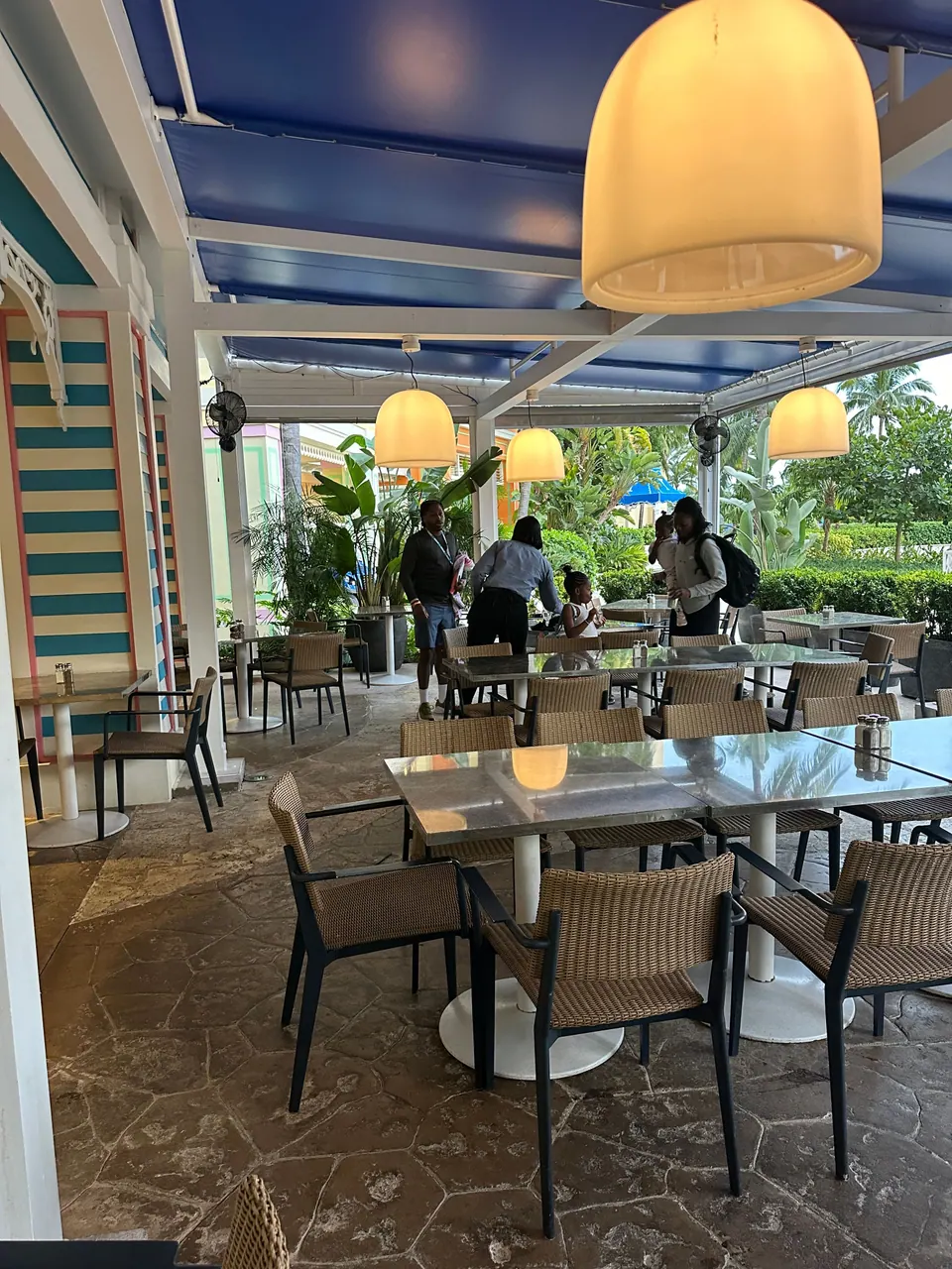 An open-air restaurant terrace with striped blue-and-white columns, woven chairs, marble-top tables and glowing pendant lamps; a few guests stand around at dusk.