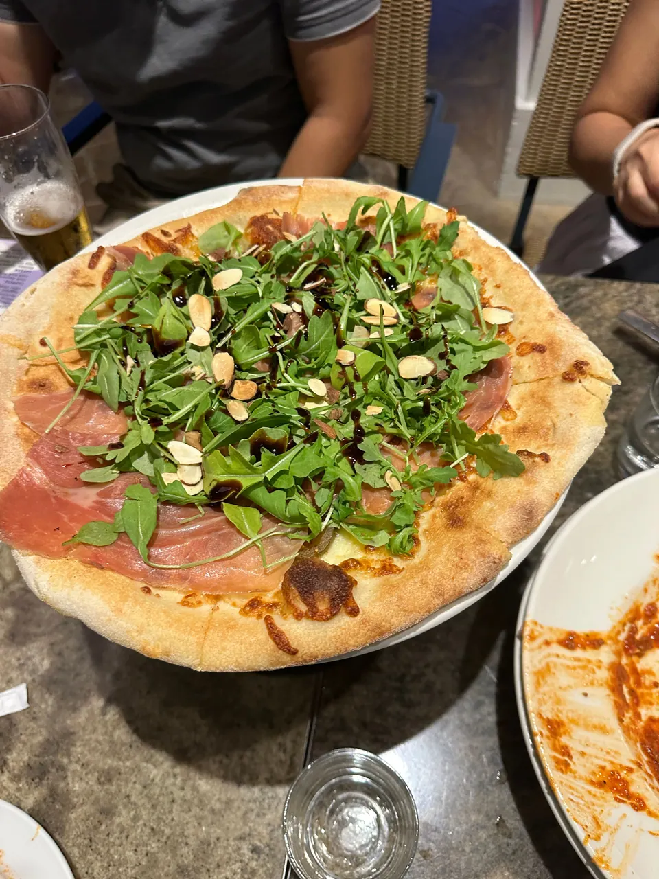 A thin-crust pizza topped with prosciutto, a heap of arugula and sliced almonds sits on a table, with a glass of beer beside it.