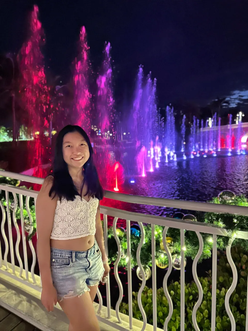 A girl in a white crochet top leans on a white scrolled railing at night while tall jets of lagoon fountains glow red, pink and purple behind her.