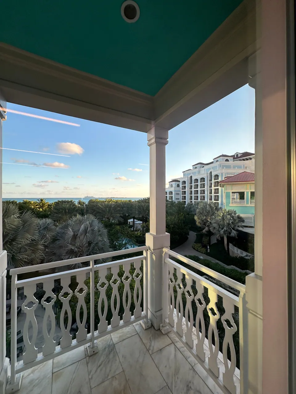 Looking out from a balcony with a white lattice railing, a low morning sky above a canopy of palms and treetops, with the resort's pastel buildings and a tower catching the early sun.