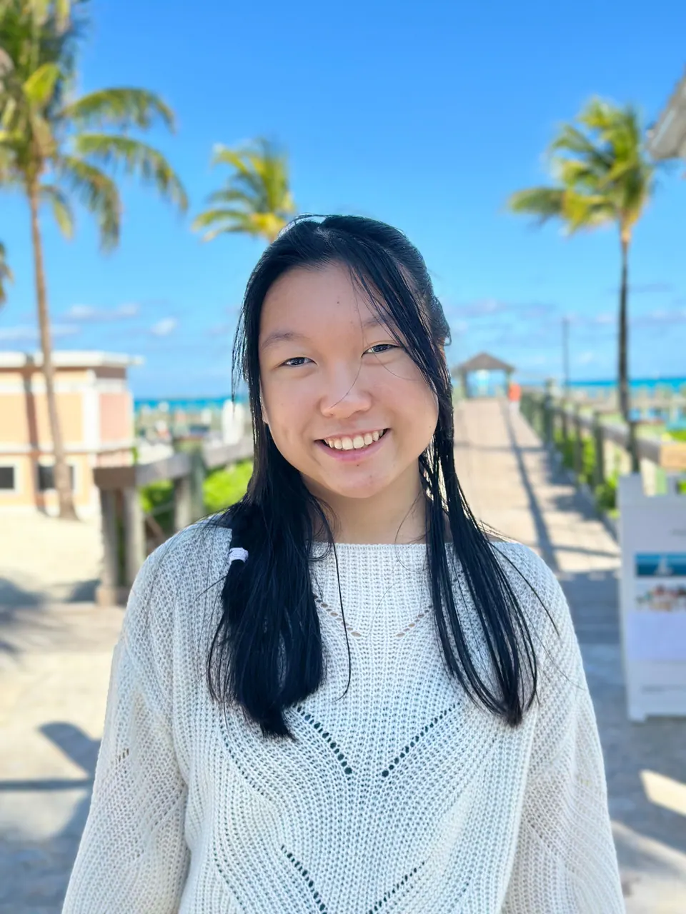 A smiling girl in a white knit sweater stands on a sunny boardwalk lined with palms that leads down to the beach.