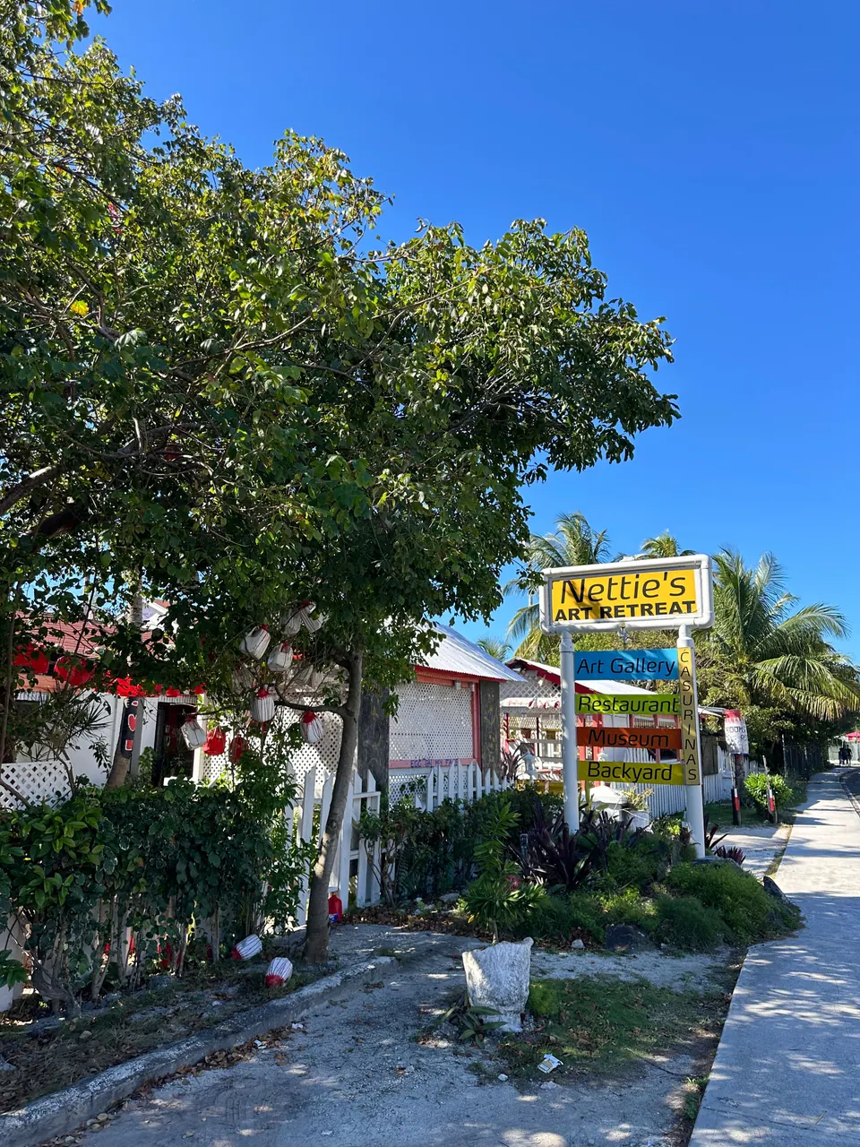 A yellow roadside sign reading Nettie's Art Retreat stands beside a white picket fence and a shady tree on a sunny, empty street of pastel cottages and palms.
