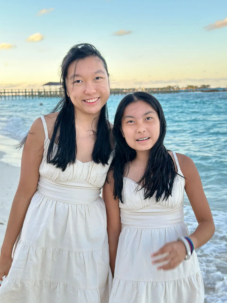 Two girls in white sundresses stand side by side in the surf with their arms at their sides, a pier and a peach and blue sky behind them.
