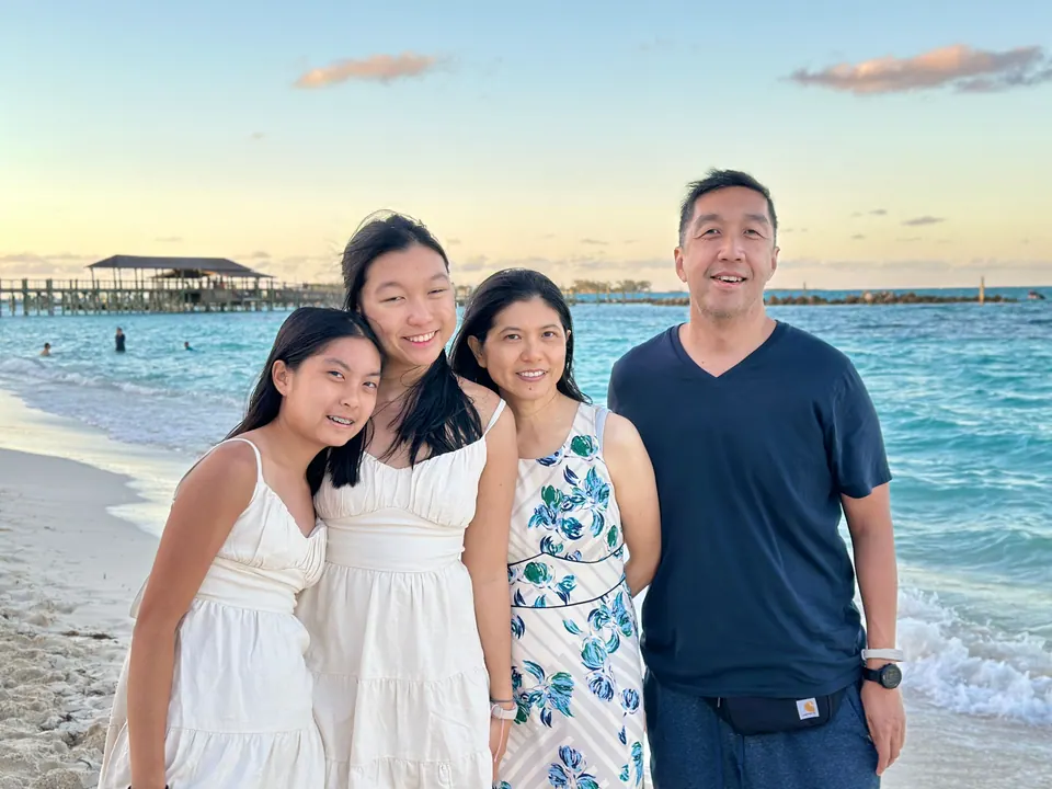 A family of four stands close together at the water's edge in golden late-afternoon light, two of them in white dresses, with a pier and calm turquoise water behind them.