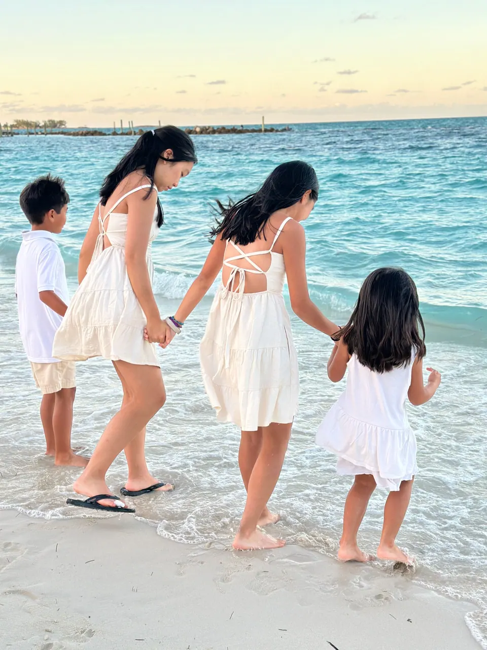Seen from behind, two girls in white dresses walk in shallow surf hand in hand with two younger children in white, all barefoot, against turquoise water.