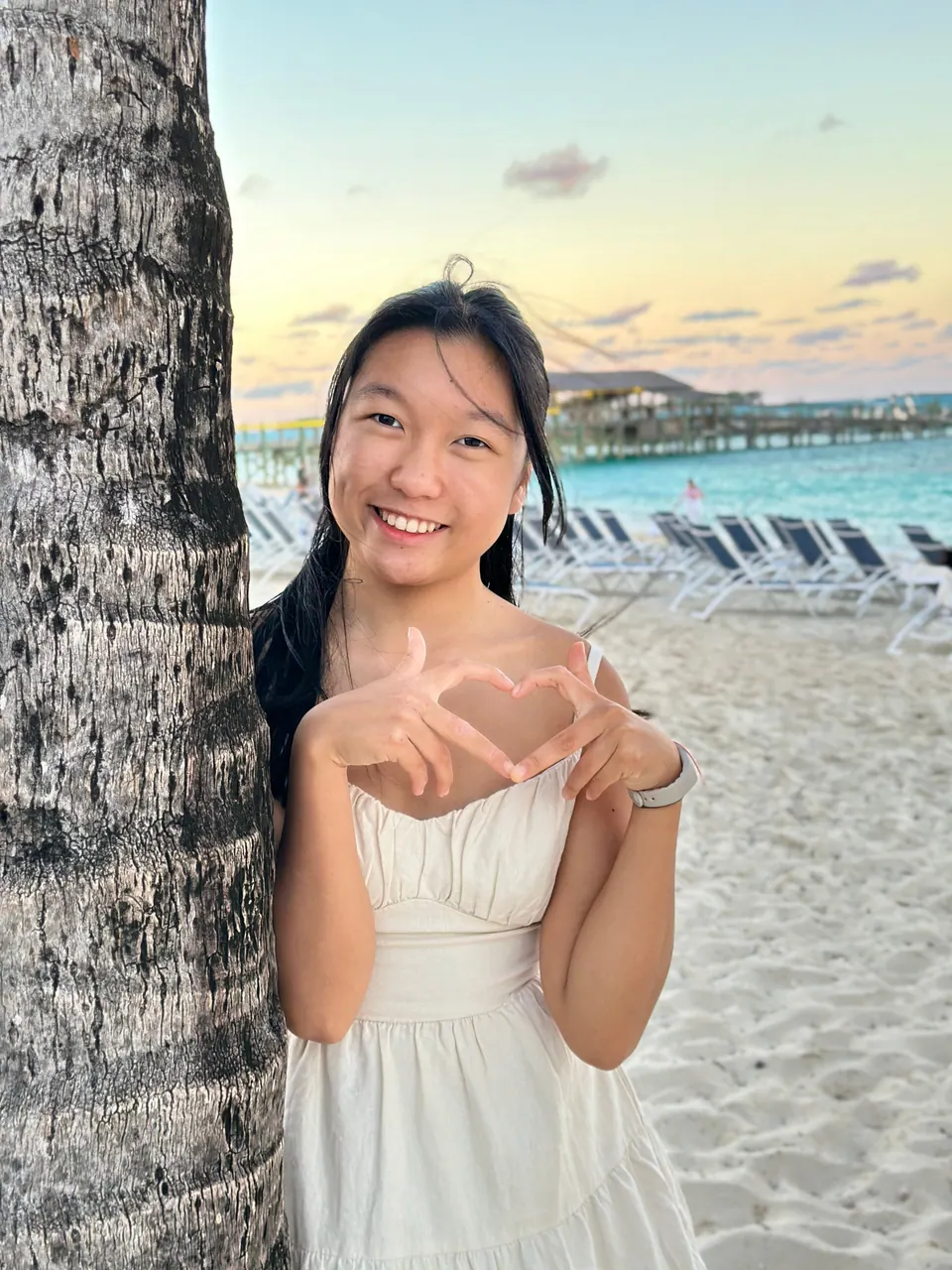 A girl leans against a palm trunk in her white dress, smiling, her hands together in a heart shape; loungers and a pier are behind her in the sunset light.