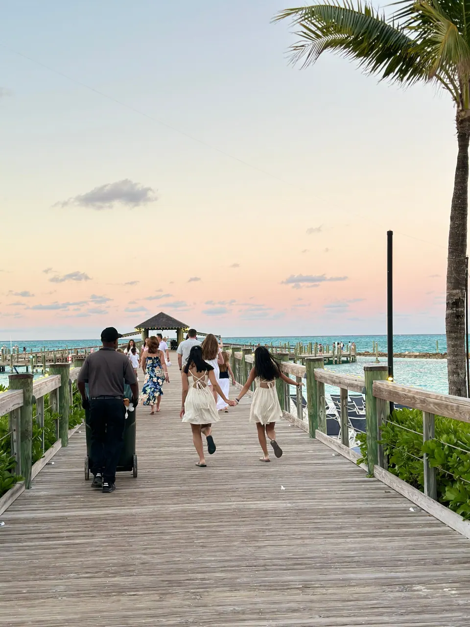 A weathered wooden boardwalk runs out toward a small gazebo over the water beneath a pastel pink and blue sky, two girls in white dresses walking hand in hand ahead of an attendant and other guests.