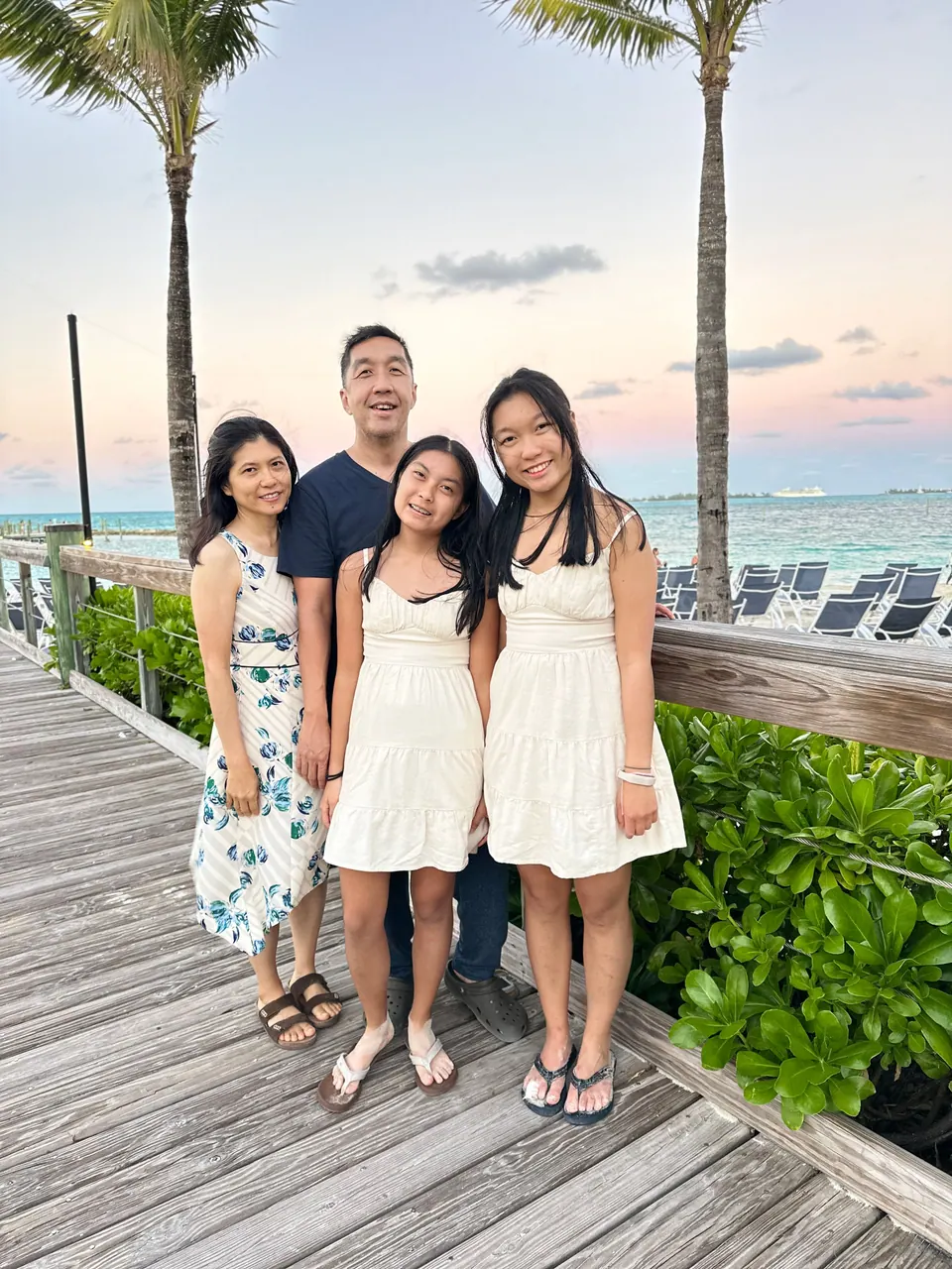 Four people stand close together on a wooden boardwalk pier, two girls in white dresses in front of a man in a navy shirt and a woman in a blue floral dress, palms and a pink evening sky behind.