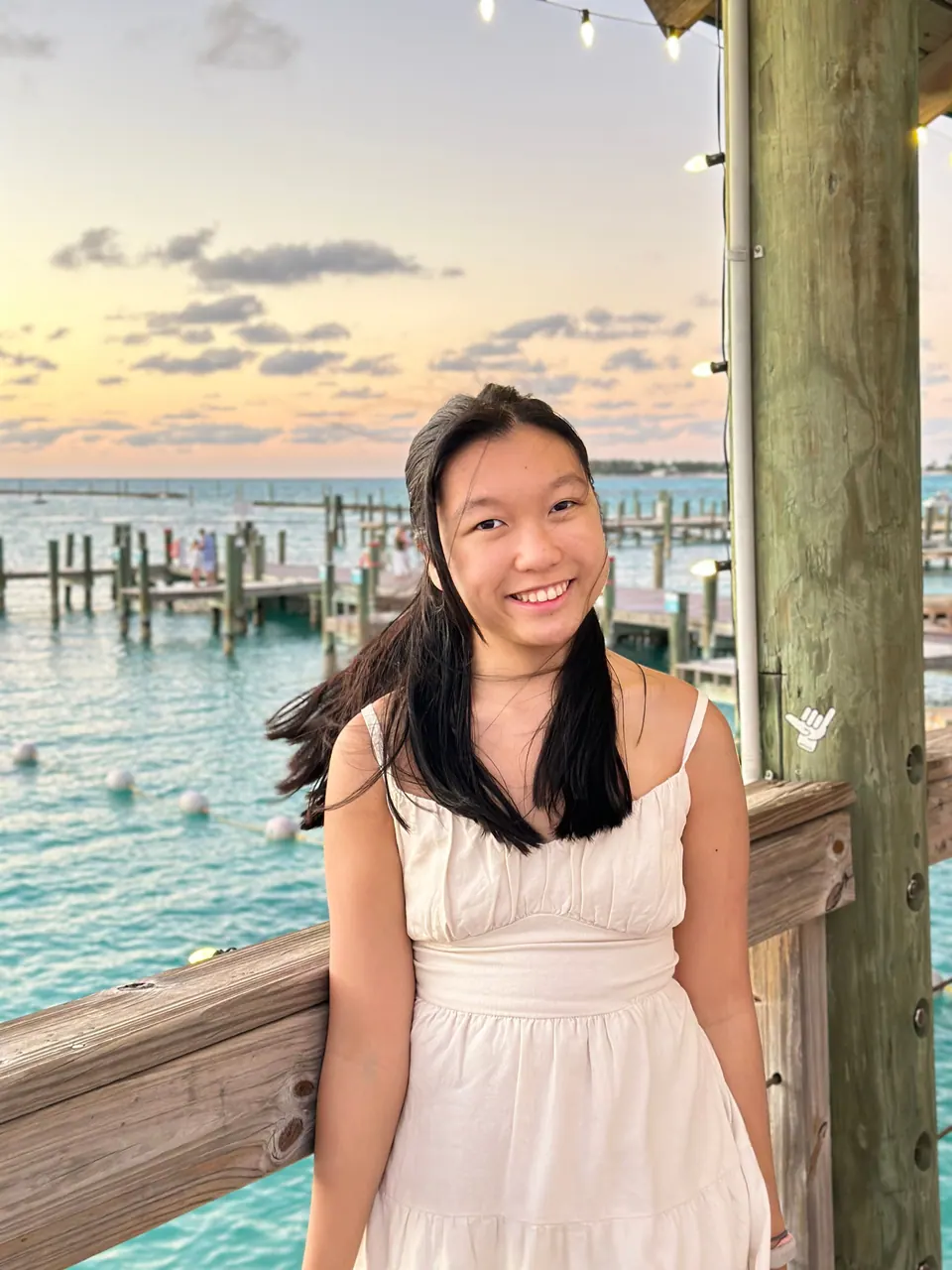 A girl in a white dress smiles, leaning on a pier railing beside a wooden post, a wooden dock, white buoys and turquoise water behind her under a golden pink sky.