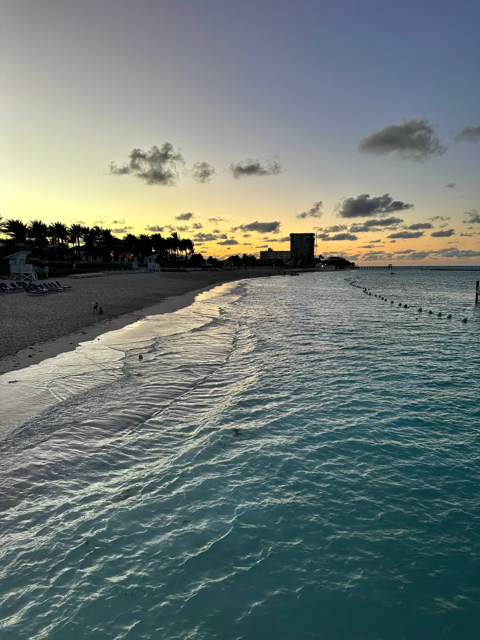 Glassy teal water curves along a pale beach toward dark palms and a hotel tower under a golden, cloud-flecked sky.