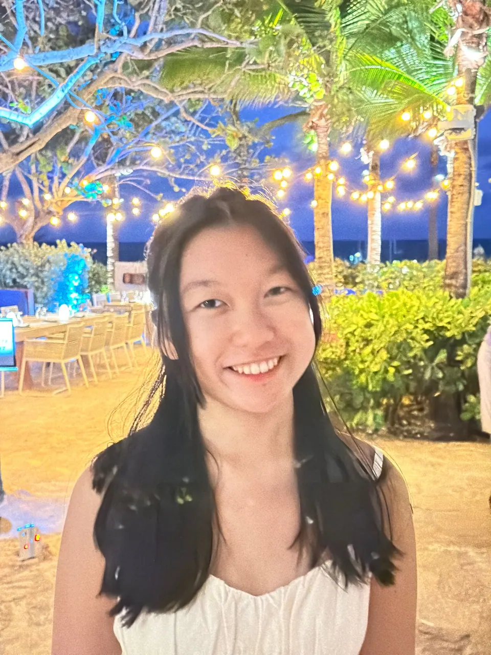 A girl in a white dress smiles into the camera with a terrace of tables, string lights and palms glowing behind her at night.