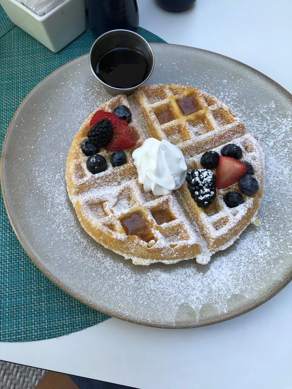 A waffle dusted with powdered sugar and topped with whipped cream, blueberries, blackberries and strawberries sits on a gray plate beside a small cup of syrup, on a teal woven placemat.