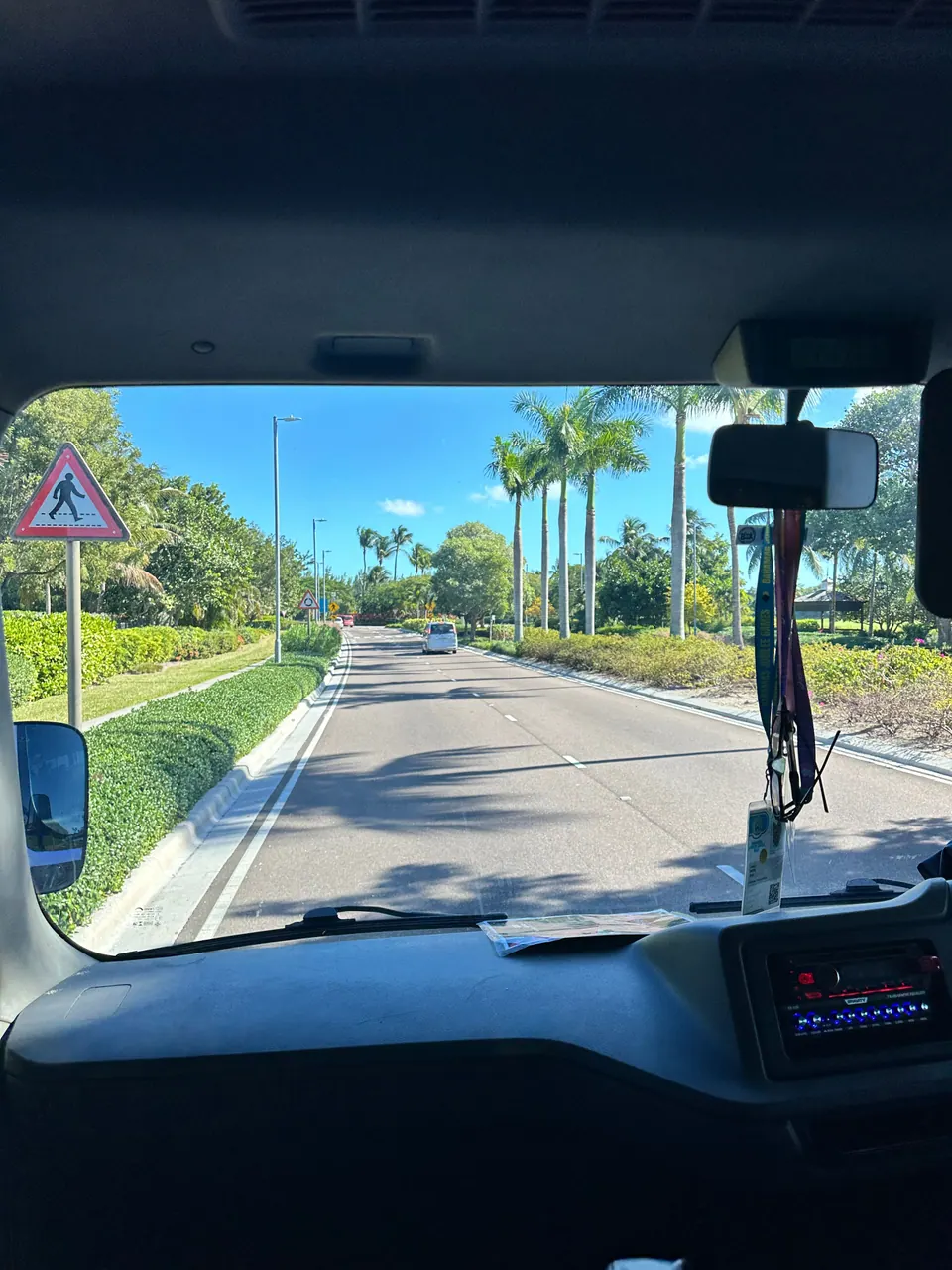 Through the front windshield of a bus, a quiet road curves between hedges and palm trees under a pedestrian-crossing sign, the mirror and dashboard in the foreground.