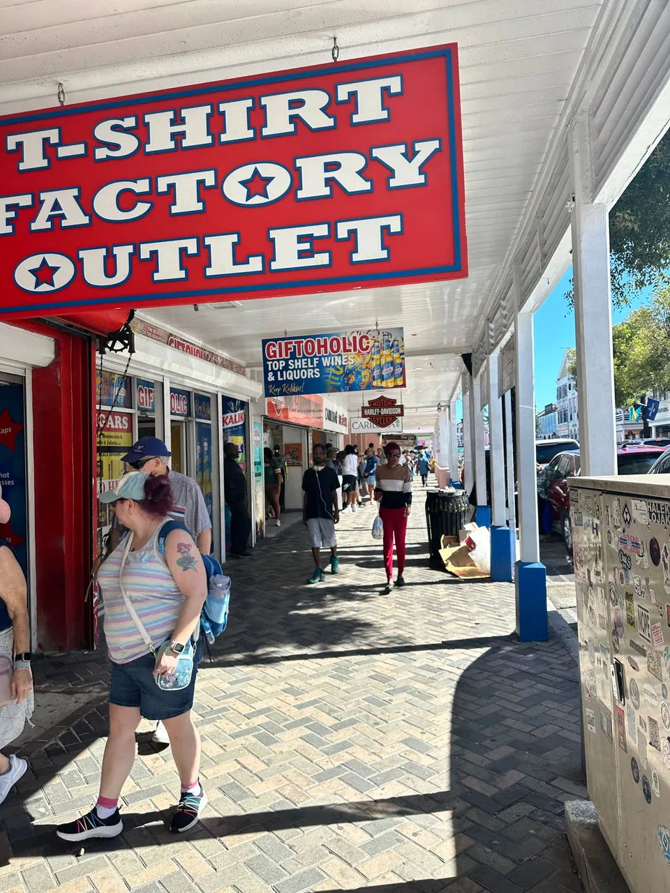 Under a white-columned arcade, a red sign reads T-Shirt Factory Outlet above shops selling souvenirs and liquor, with visitors strolling along the brick pavement.