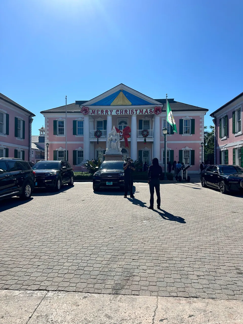 A pink Georgian government building with a blue and yellow pediment, a green flag and a Merry Christmas banner faces a brick square with a statue in front and dark SUVs parked to either side.