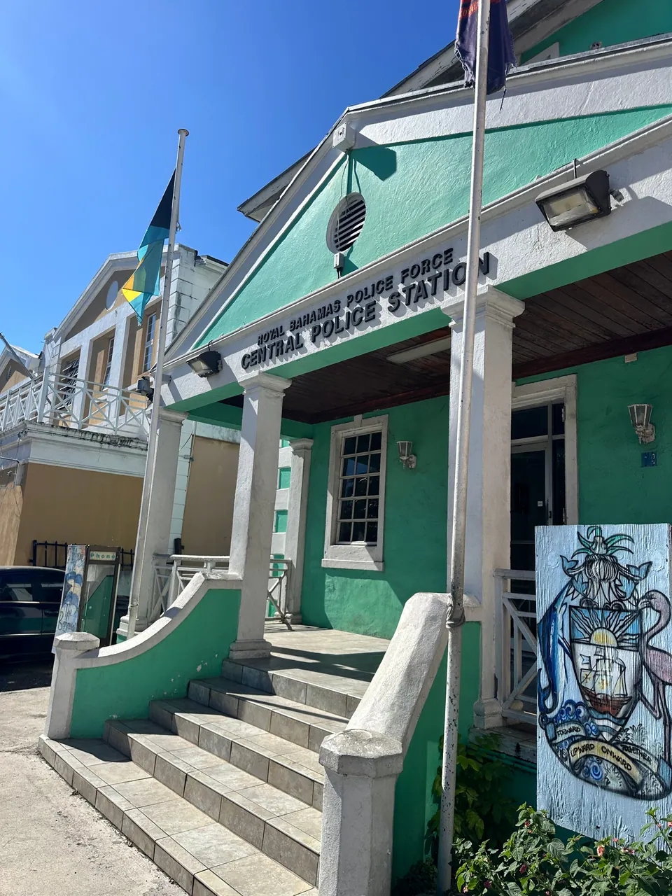 A turquoise and white colonial building with a columned porch and steps carries a sign reading Royal Bahamas Police Force, Central Police Station, with a Bahamian flag and a coat-of-arms mural at the side.