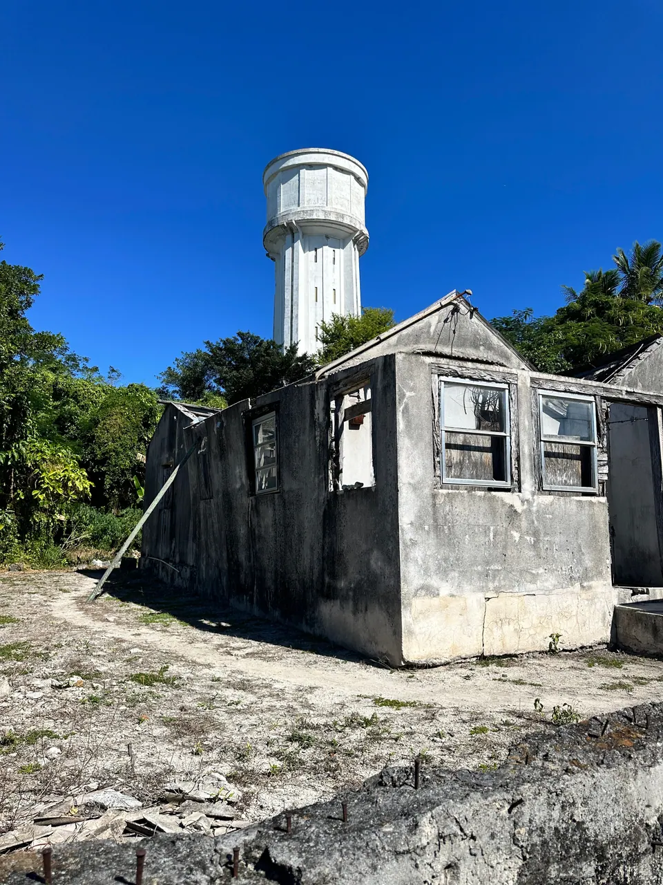 A weathered gray stone building with empty window openings and a broken roof stands on bare ground, a tall white cylindrical water tower rising behind it against a deep blue sky.