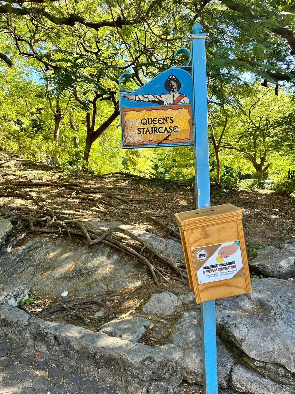 A hand-painted Queen's Staircase sign on a blue post, with a yellow donation box beneath it, stands among tree roots and rock in dappled sun.