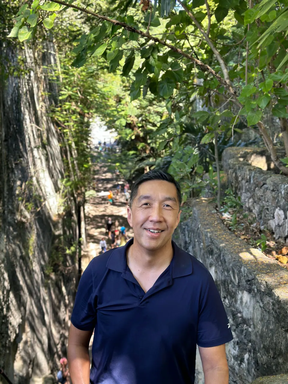 A smiling man in a navy polo shirt poses in the shade of a limestone gorge with its stone wall, trees and a few visitors behind him.