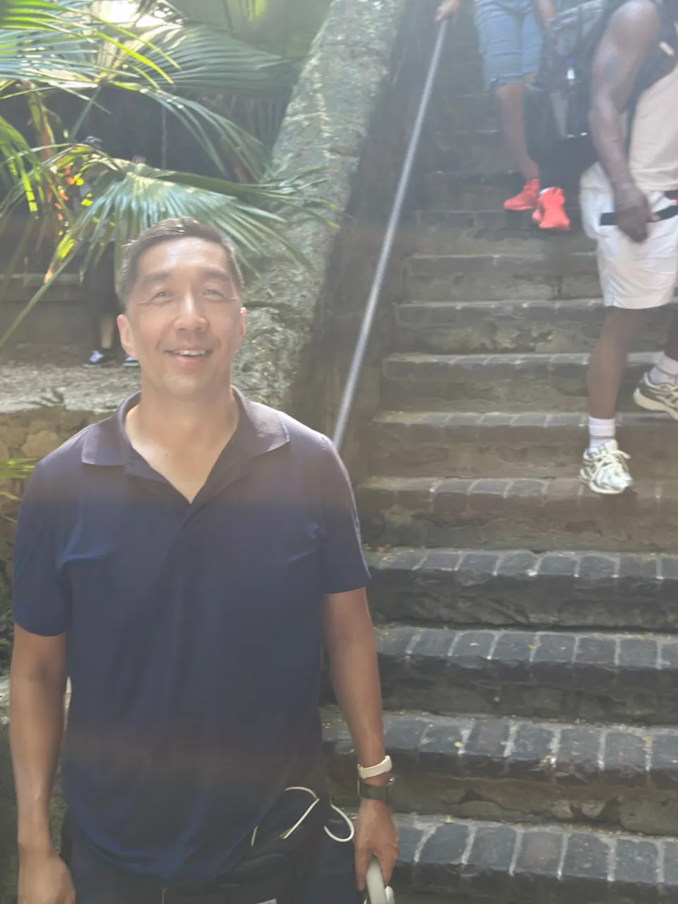 A man in a navy polo stands on worn stone steps beside a metal handrail as other visitors climb behind him.