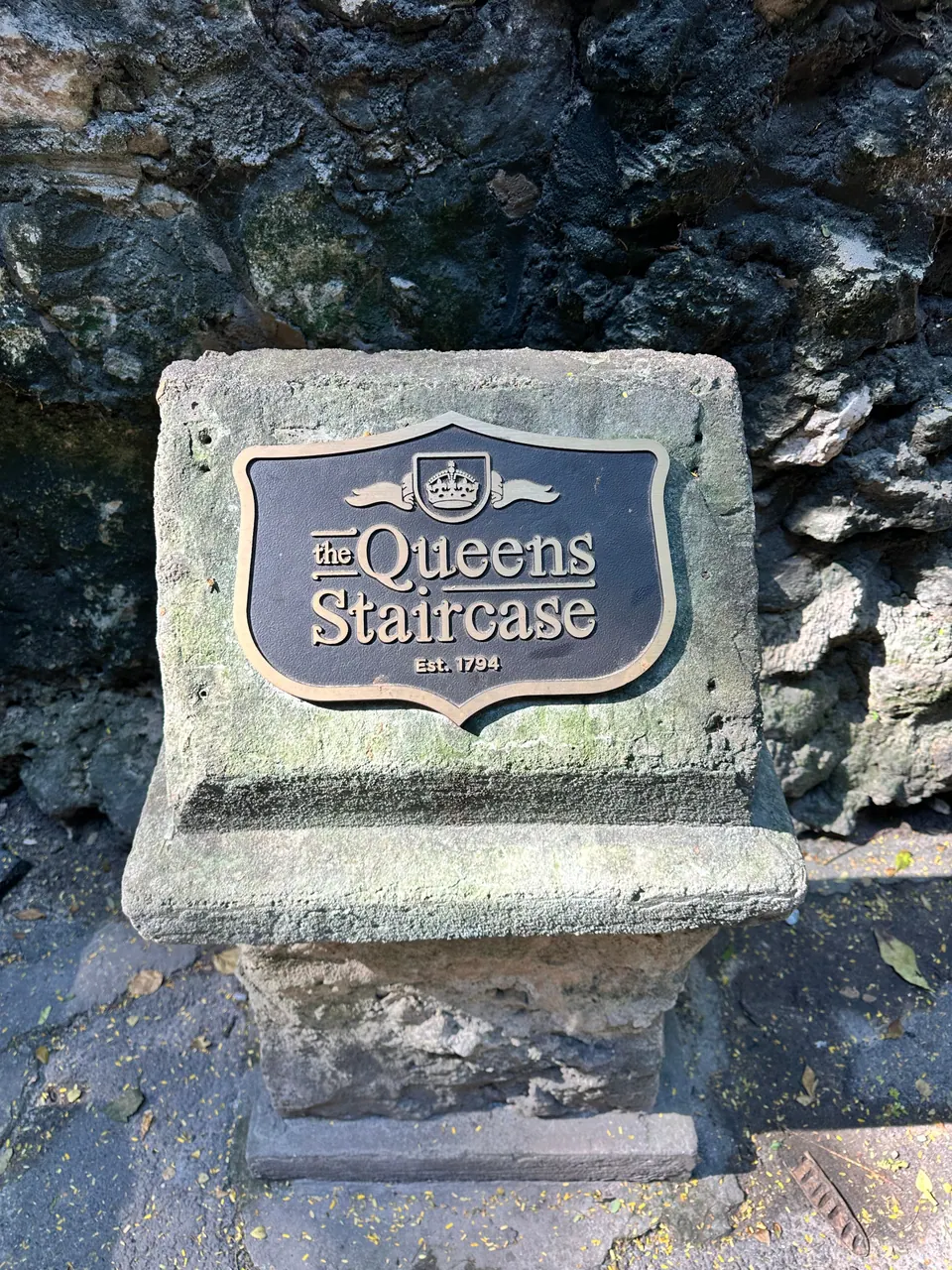 A bronze shield-shaped plaque reading The Queen's Staircase, Est. 1794, sits on a stone pedestal against dark rock.