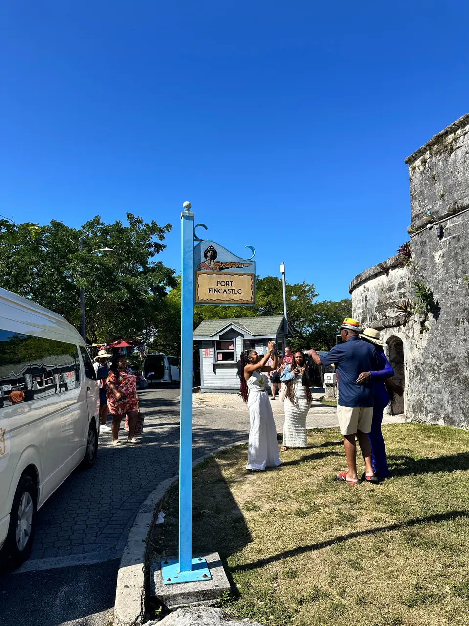 A blue post carries a Fort Fincastle sign beside a white van, with visitors posing for photographs on the grass in front of the stone fort wall.