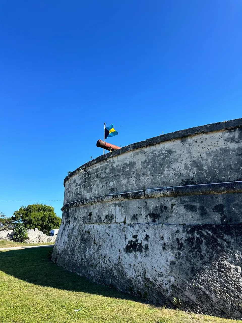 The curved whitewashed stone wall of the fort rises above a lawn, topped by a Bahamian flag and a cannon, against a cloudless blue sky.