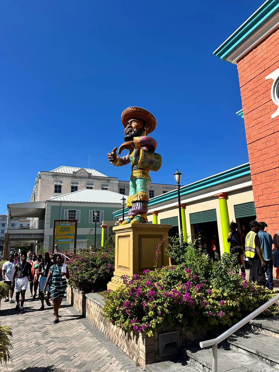 A brightly painted statue of a figure in a wide hat stands on a plinth among flowers outside an orange and green building, people walking past under a deep blue sky.