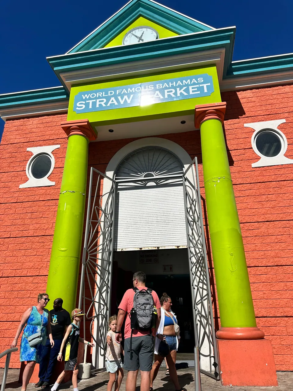 The orange-red front of a market building has lime-green columns, porthole windows and a sign reading World Famous Bahamas Straw Market, with visitors coming and going through the arched door.