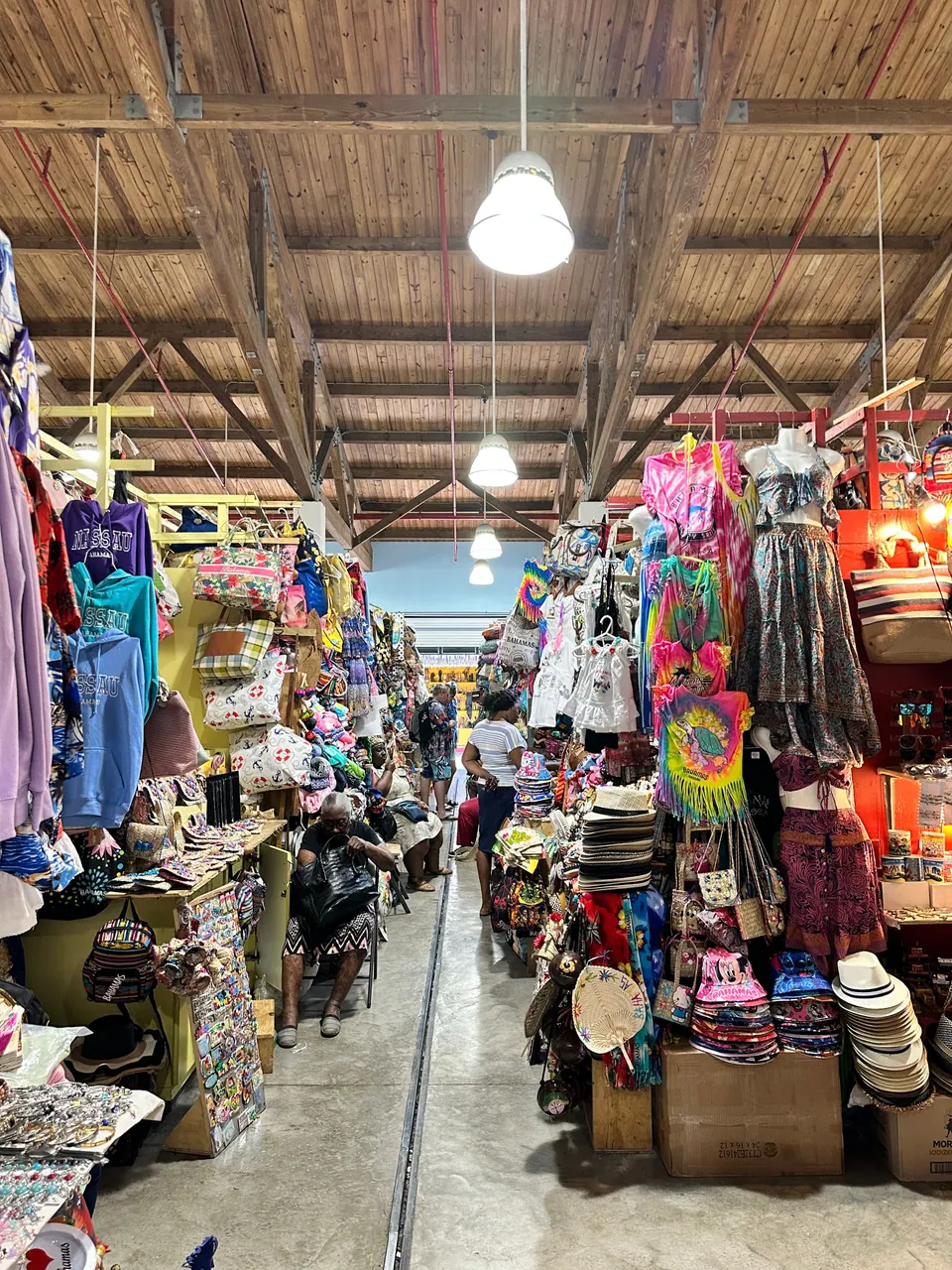 A long aisle inside a market hall under wooden rafters, lined on both sides with stalls of hats, woven bags, beach dresses and T-shirts.