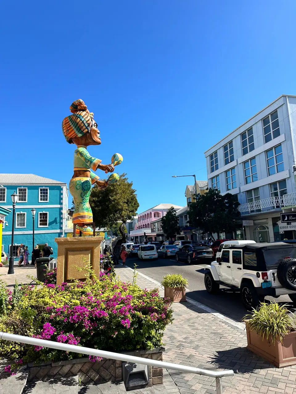 A brightly painted statue of a woman in a turban holding maracas stands on a plinth above a flower bed, beside a turquoise building and a street with a white Jeep.