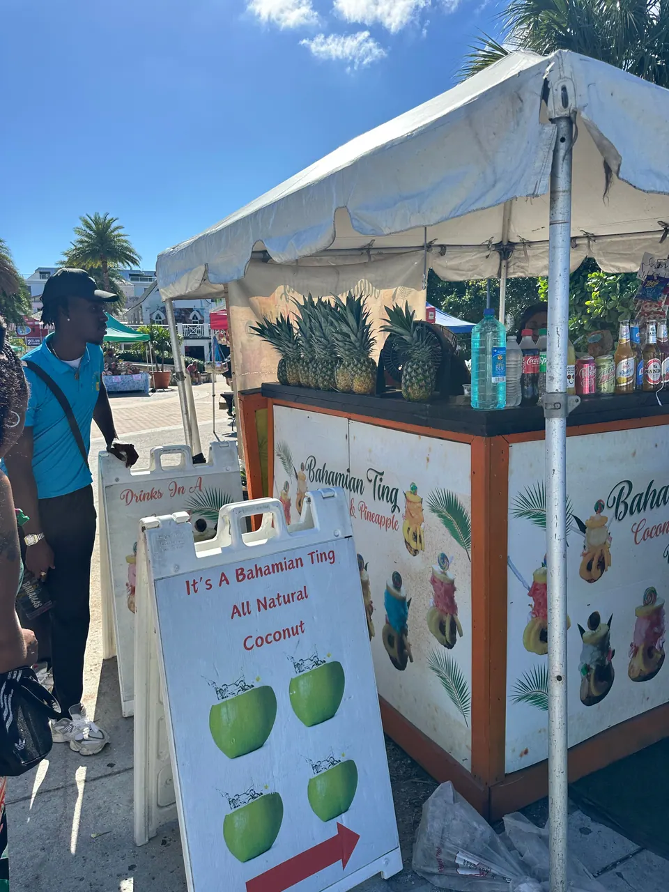 A white-canopied juice stand piled with pineapples has a folding sign reading It's a Bahamian Ting, All Natural Coconut, and a vendor at its side.