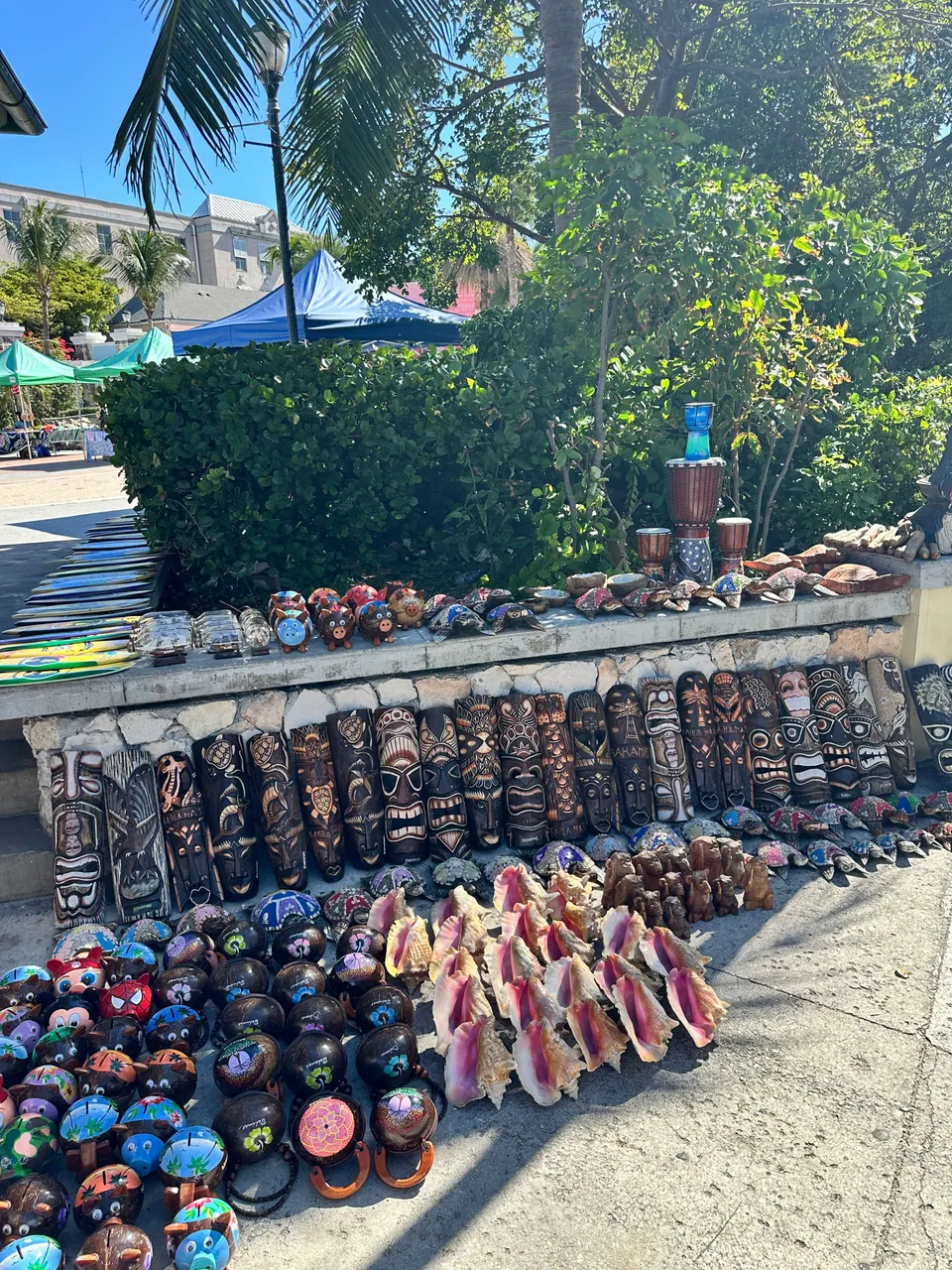 Rows of carved wooden masks, painted gourd bowls and pink conch shells are laid out along a low wall at an open-air craft stall beside a hedge and tents.