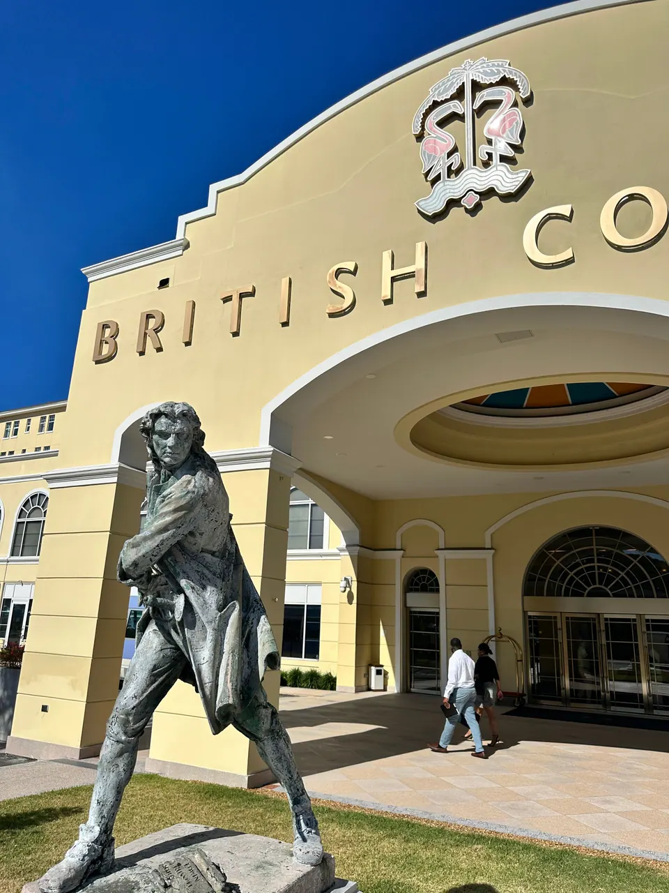 A dark bronze statue of a man in an eighteenth-century coat stands on the lawn in front of the cream-coloured arched entrance of a hotel, with a couple walking past.