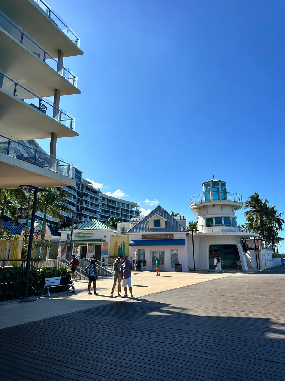 A paved waterfront plaza of pastel shops with a small white lookout tower and a glass hotel tower beside it, under a bright blue sky.