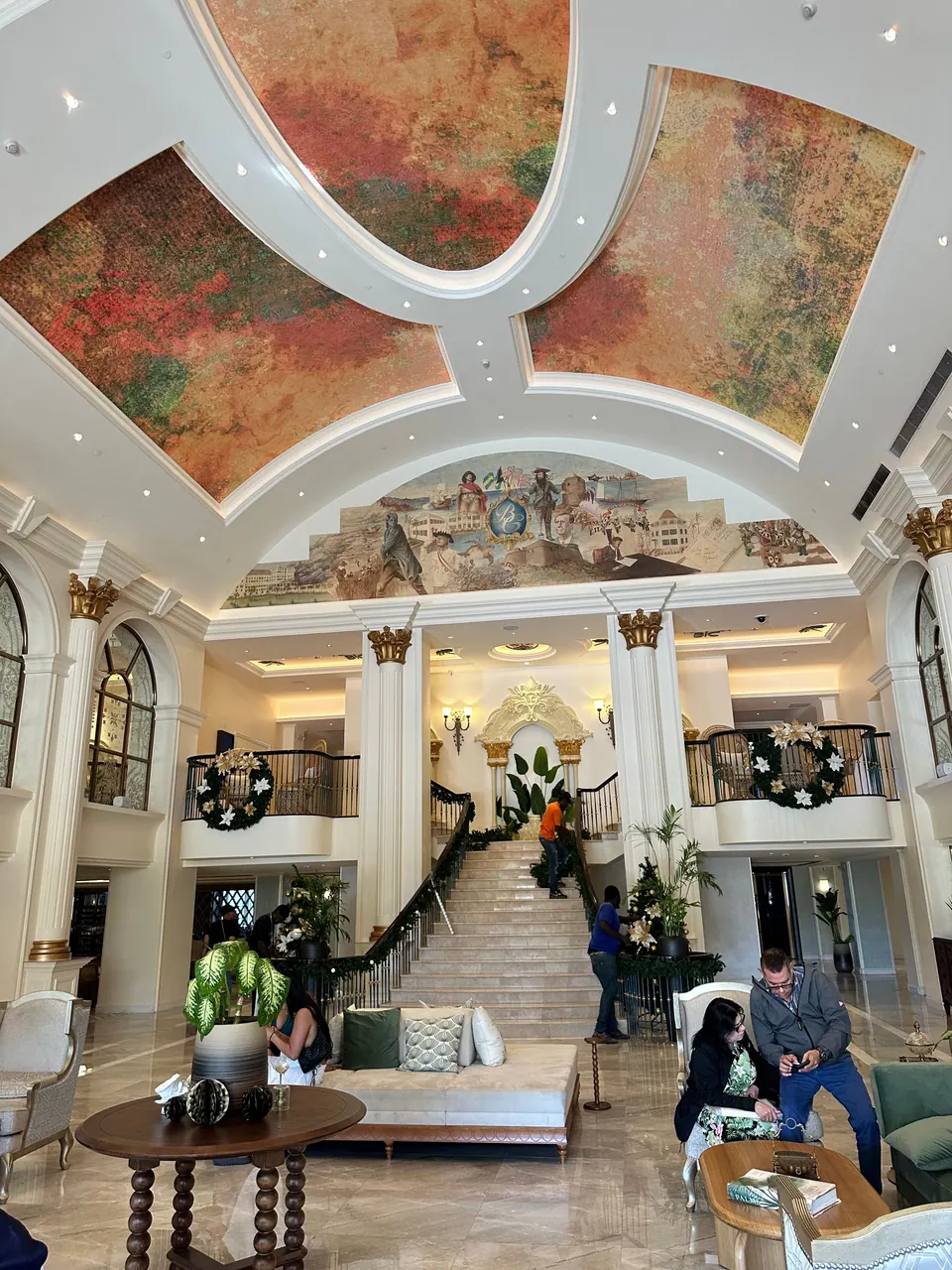 A high hotel lobby with a painted ceiling and a large mural above a double staircase, wreaths on the balconies and guests seated in armchairs below.