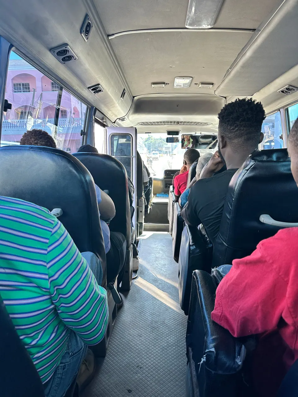 Inside a bus, passengers in black high-backed seats face forward down the narrow aisle; through the windows pink buildings show on the street.