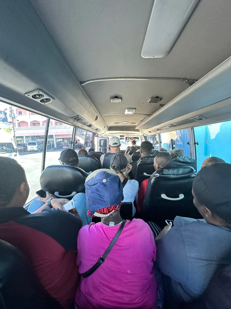 Looking down the length of a full bus, rows of passengers sit and stand in the aisle beneath the ceiling vents, a girl in a pink top and cap in the foreground.