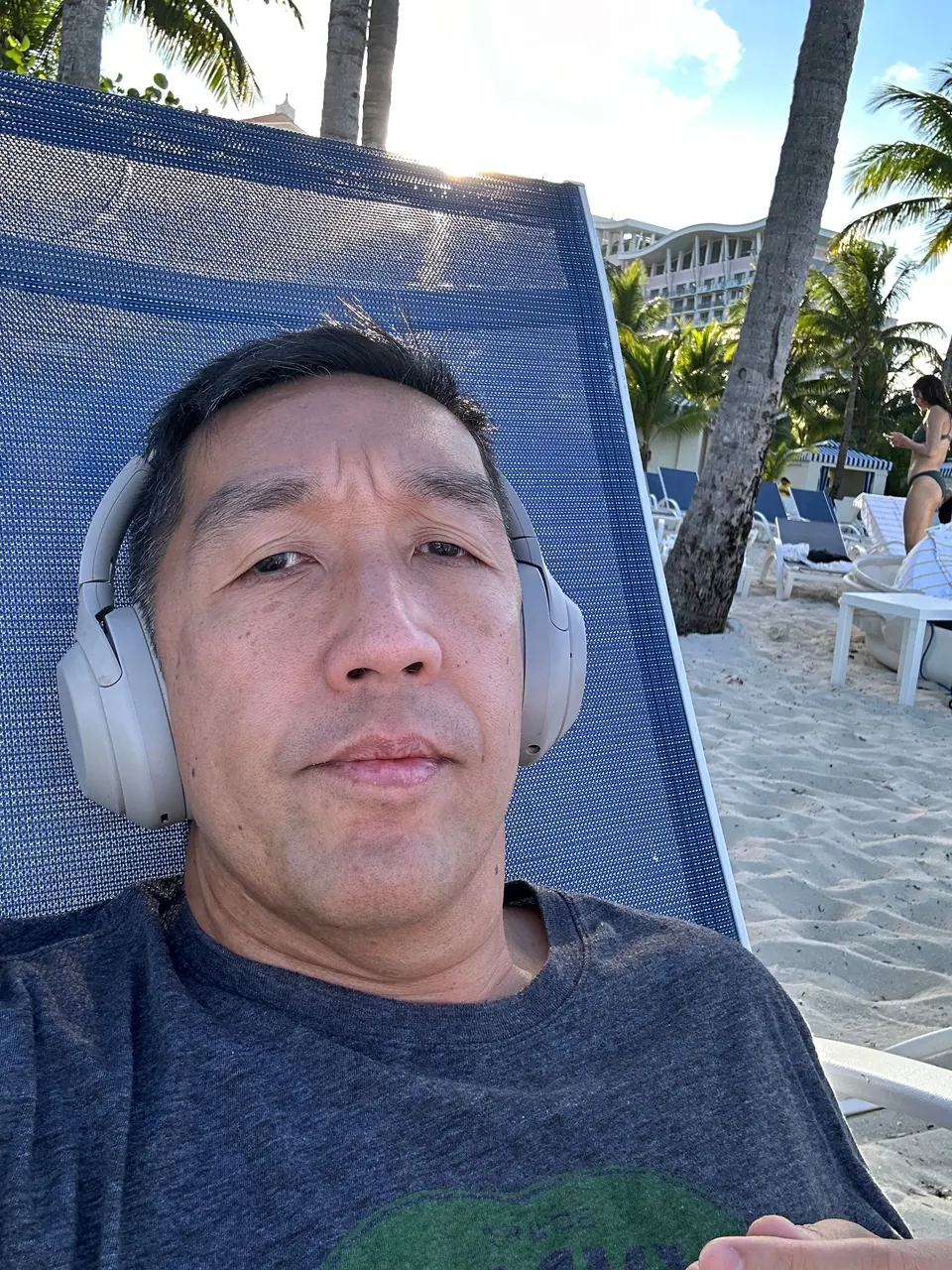 A man in gray over-ear headphones takes a selfie reclined on a blue beach lounger with white sand and palm trees behind him.