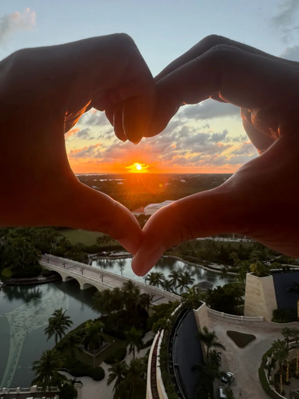 Two hands make a heart shape framing a low orange sun and clouds above a green landscape, with a white arched bridge, a lagoon and resort paths far below.
