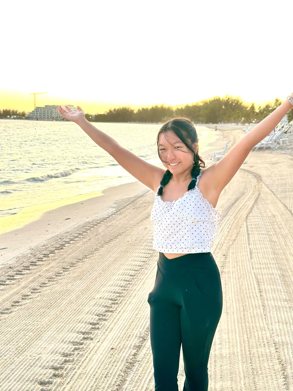 A girl in two braids, a white polka-dot top and black trousers throws her arms wide and smiles on the sand, the low sun glowing over the water and a hotel on the shore behind her.