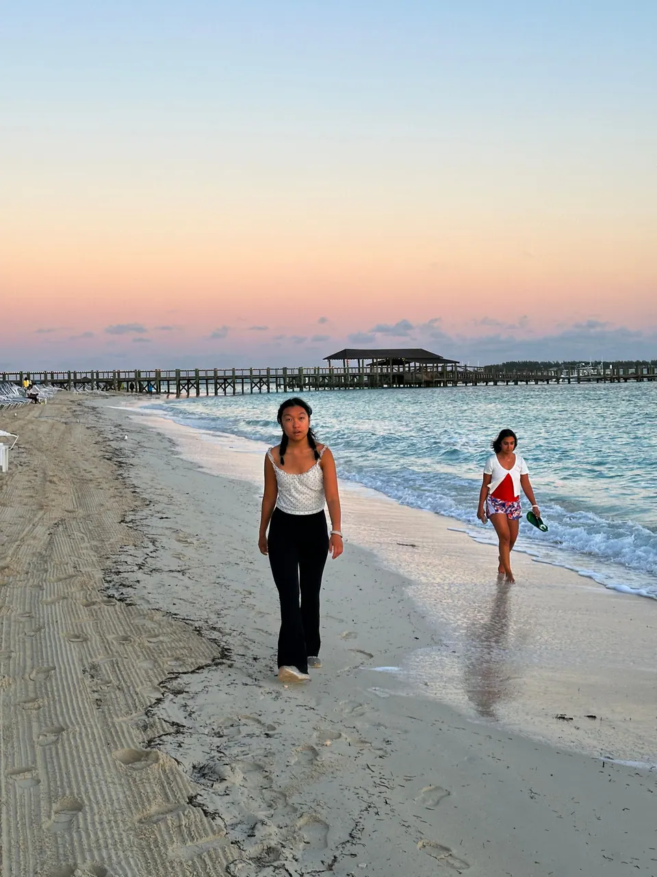 A girl walks toward the camera along the wet sand at the water's edge under a pink and orange dawn sky, the beach pier and gazebo in the distance and another walker at the right.