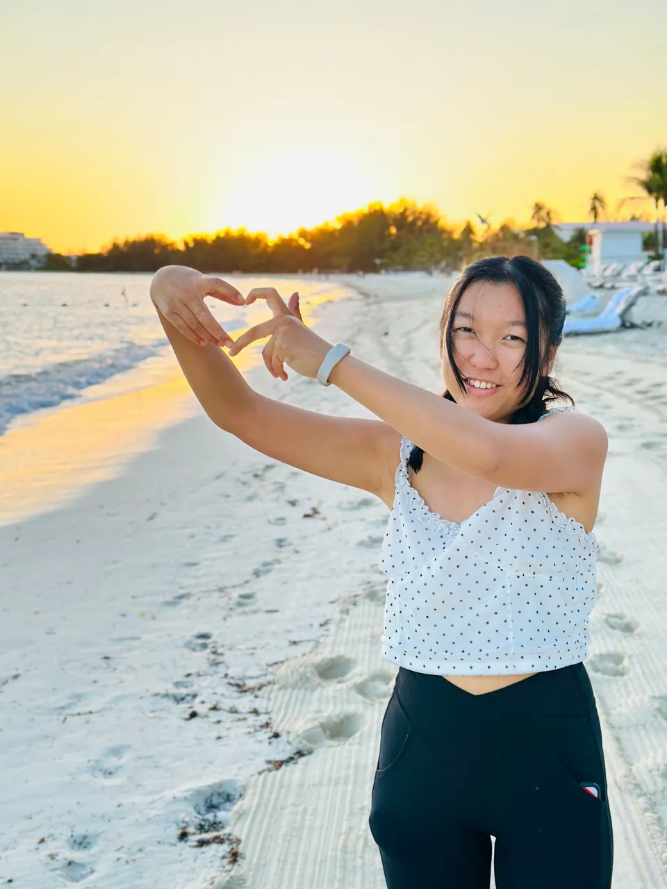 A girl makes a heart shape with her hands and smiles at the camera on the pale sand, the sun rising gold behind a line of trees and lit surf at the left.