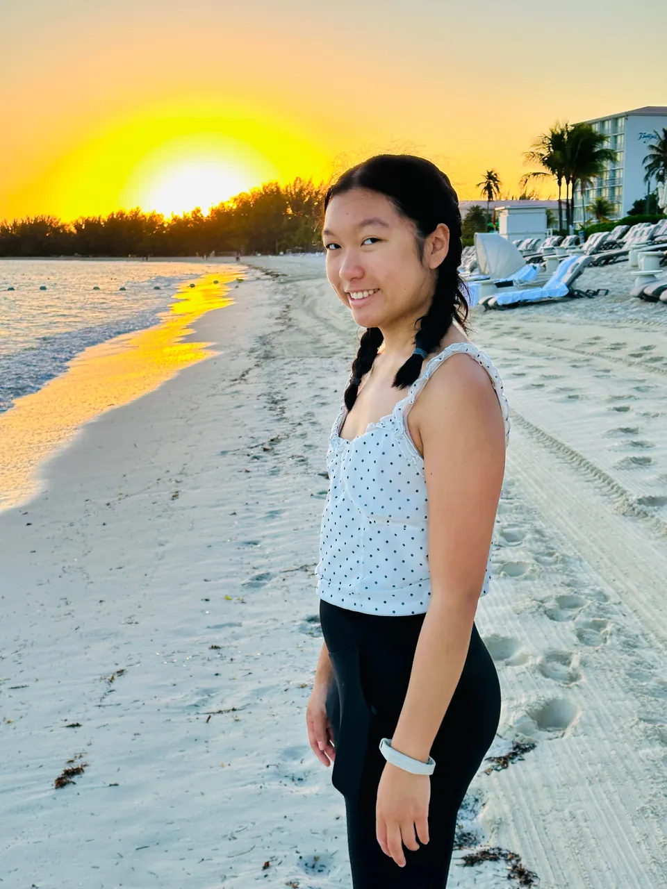 A girl with her hair in two braids smiles back at the camera on the beach, the sun rising as a golden ball behind her and loungers along the sand.