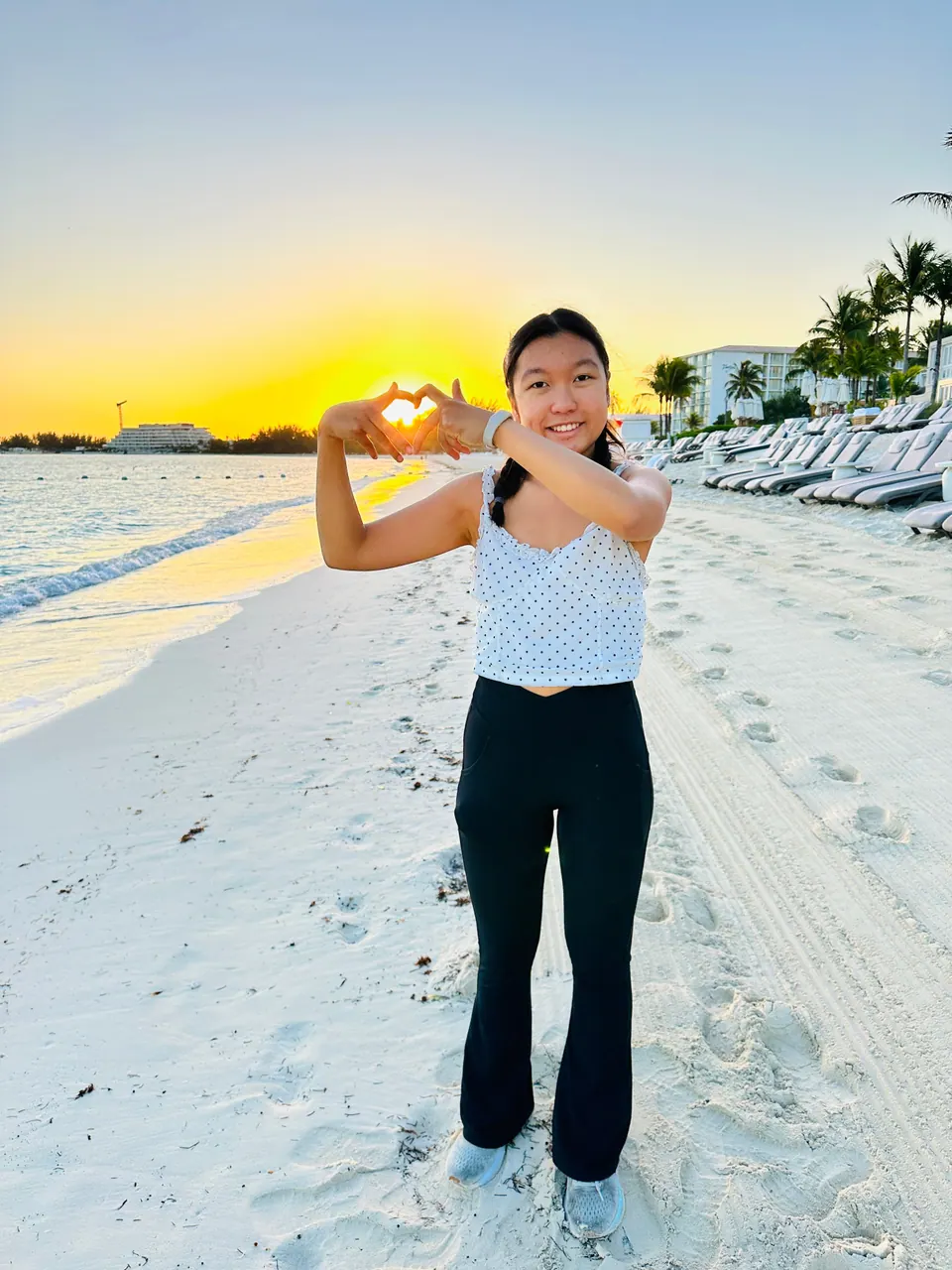 A girl stands on the sand with her hands forming a heart around the low, glowing sun, loungers and palms along the beach behind her.