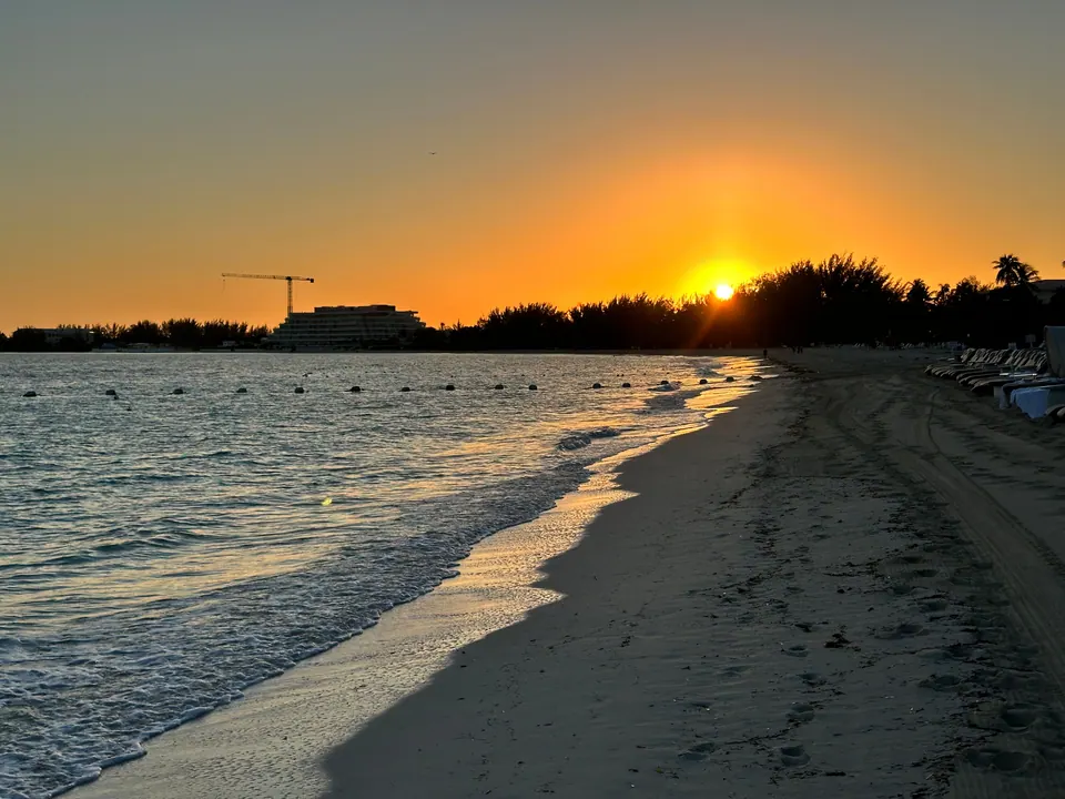 The sun sits just above a line of trees and a construction crane, the orange sky reflected on wet sand and gentle surf curving away along the beach.