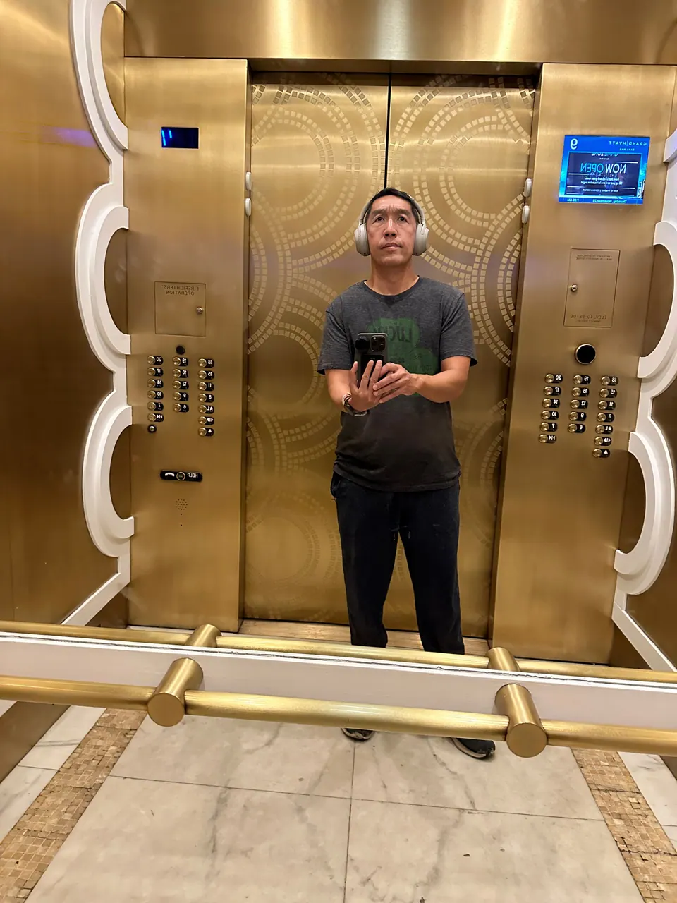 A man in headphones and a gray T-shirt photographs himself in the mirrored gold doors of a hotel elevator, phone in hand.