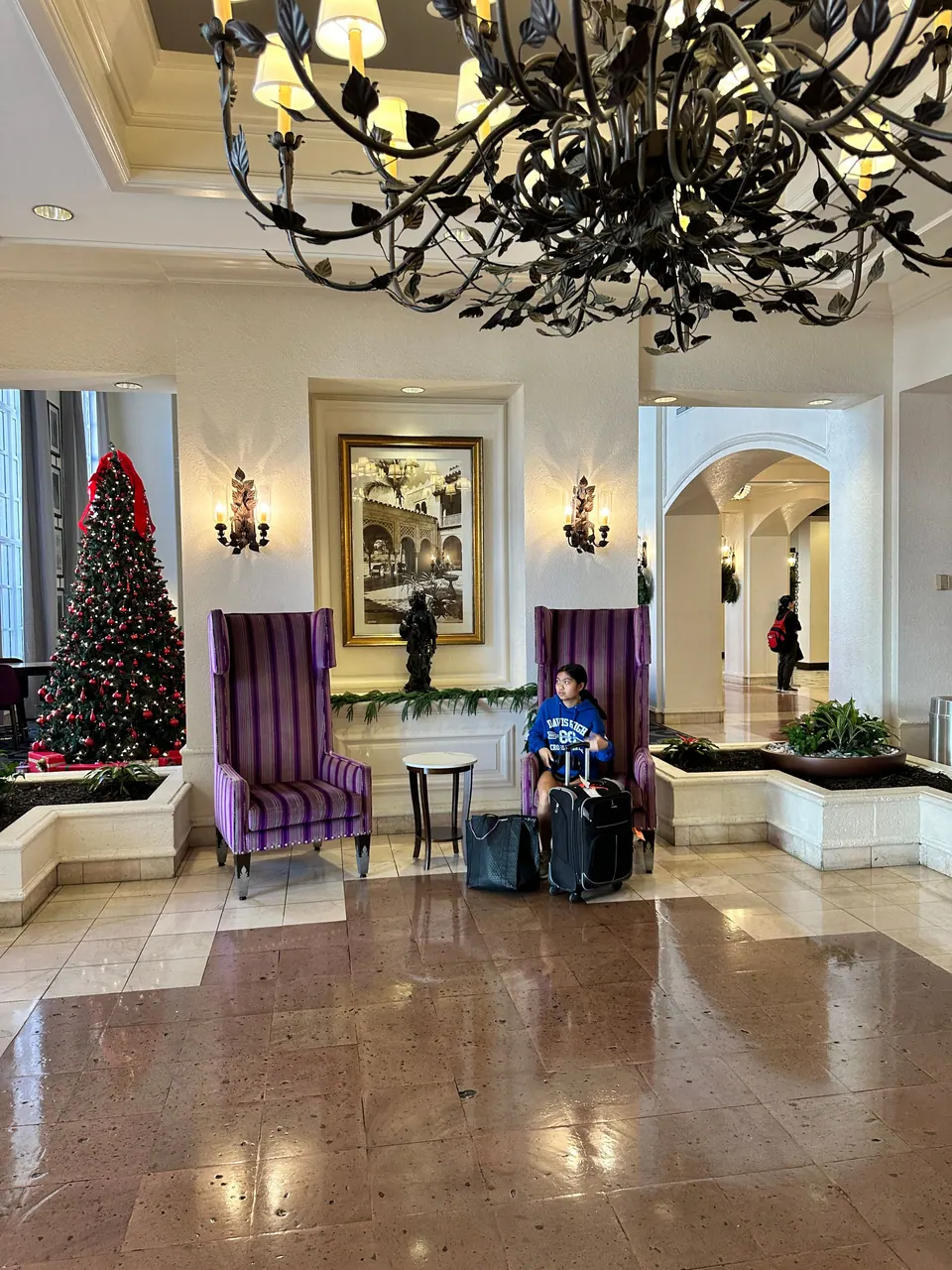 A girl sits with suitcases between two tall purple chairs in a marble hotel lobby under a wrought-iron chandelier, a Christmas tree to the left.