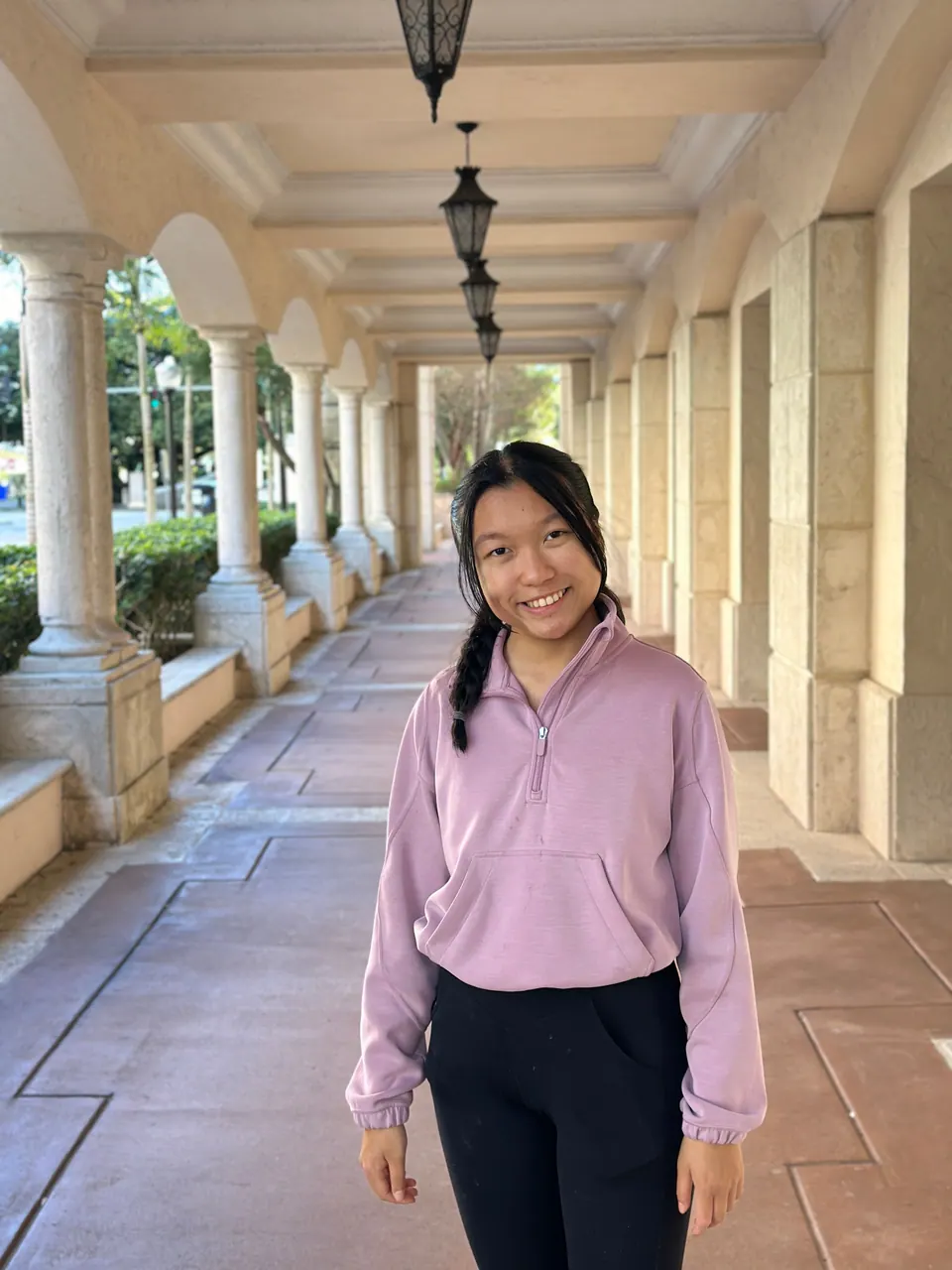 A girl in a lilac zip-neck pullover and black trousers, her hair in two braids, smiles in a long shaded arcade of cream columns with hanging lanterns.